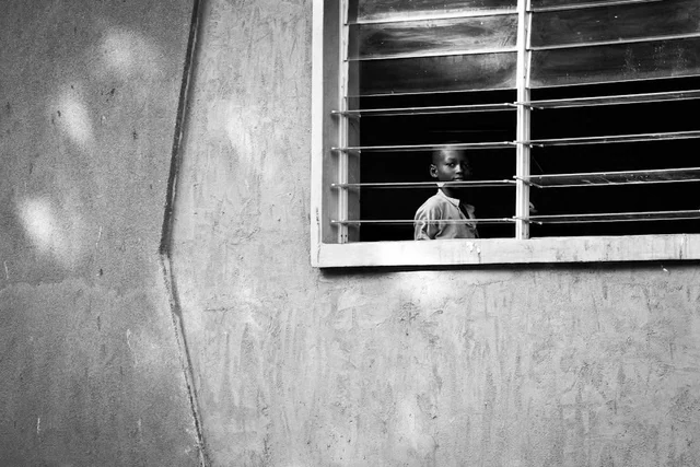 A child looks out from behind the horizontal bars of a window in a wall patterned with patches of light, in black and white.