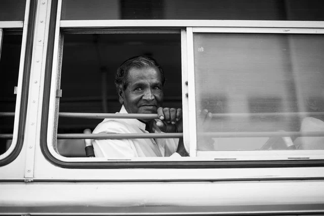 Um passageiro olha para o fotógrafo através de uma janela aberta com barras horizontais, a preto e branco.