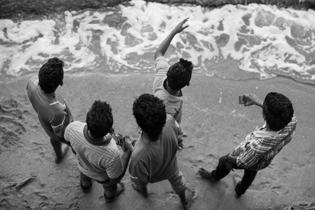 Cinco homens vistos de cima à beira de água, um deles a levantar o braço diante das ondas, a preto e branco.