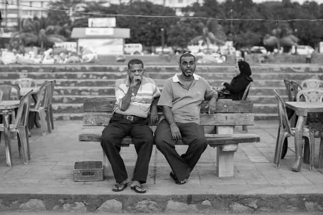 Dois homens sentados num banco de madeira diante de degraus e de uma esplanada, fotografados a preto e branco.