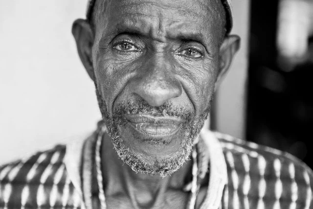 Retrato de frente a preto e branco de um homem com barba curta grisalha, vestido com uma camisa estampada.