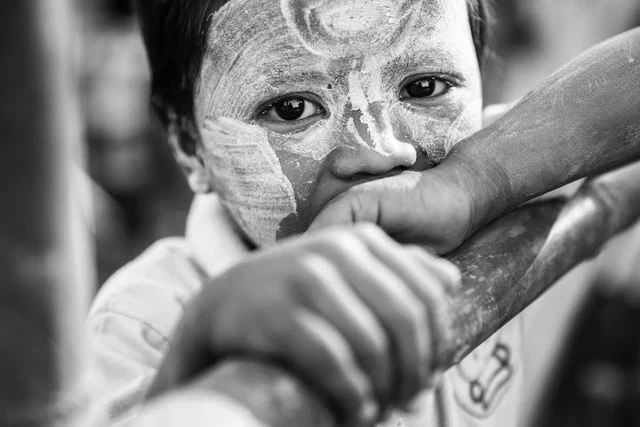 Retrato a preto e branco de uma criança com o rosto coberto de desenhos claros e os braços apoiados numa barra.