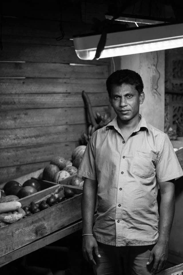 Retrato a preto e branco de um homem de pé ao lado de caixas de legumes numa pequena loja.