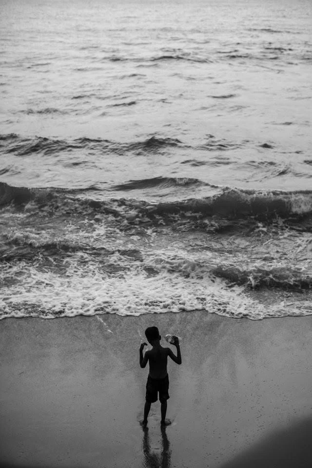 Uma criança vista de costas na areia, de frente para as ondas, fotografada de cima a preto e branco.