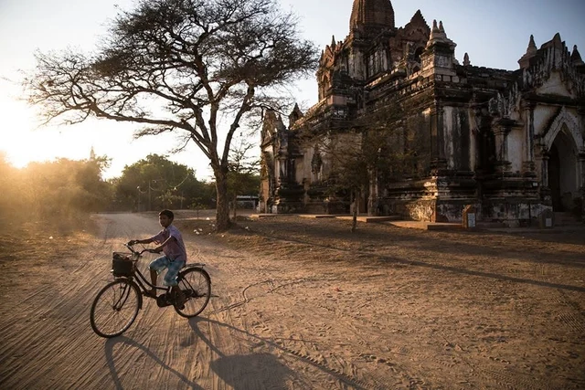 A child rides a bicycle past a temple and a large tree in golden light casting long shadows.