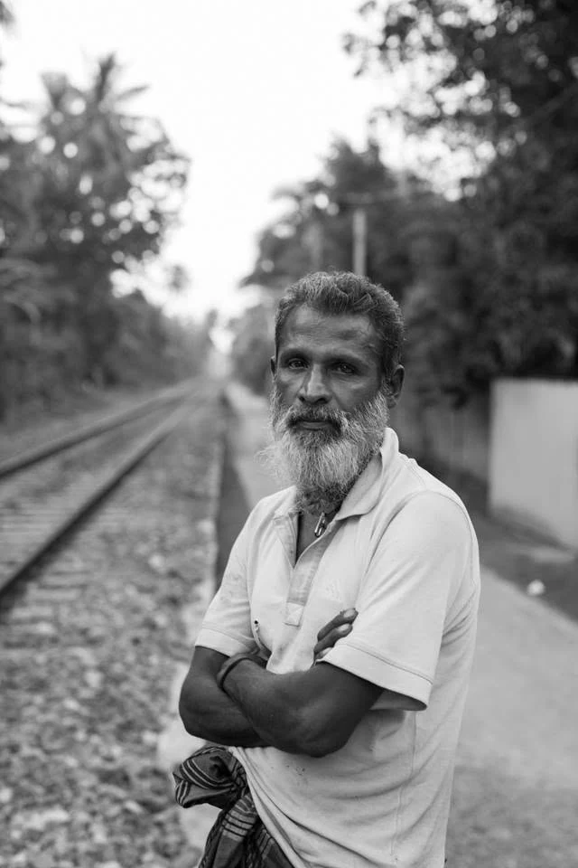 Retrato a preto e branco de um homem de barba branca, de braços cruzados junto a uma linha férrea.