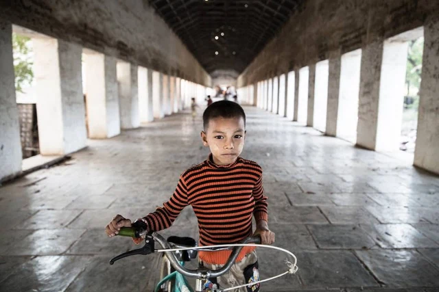 Uma criança com uma camisola às riscas vermelhas e pretas segura uma bicicleta numa longa galeria ladeada de colunas.