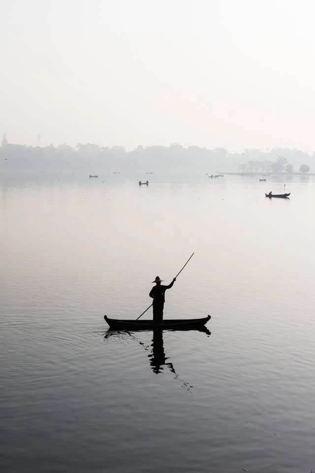 A silhueta de uma pessoa de pé num barco reflete-se na água calma, com outras embarcações na neblina.