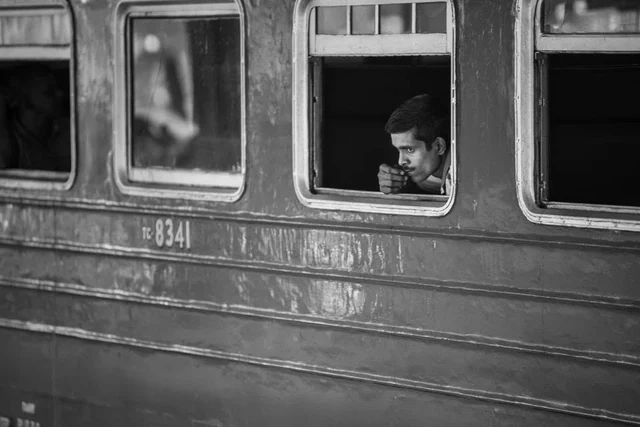 A man looks out through the open window of an old railway carriage, in black and white.