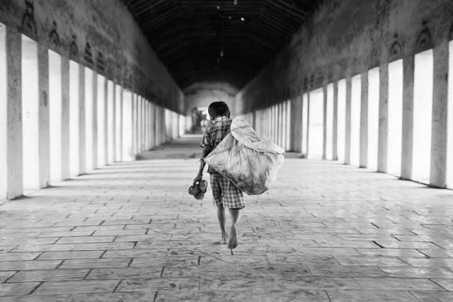 Uma pessoa caminha descalça numa longa galeria com colunas, transportando um grande saco e os sapatos na mão, a preto e branco.