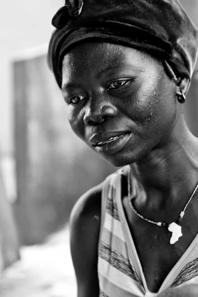 Retrato a preto e branco de uma mulher com um lenço escuro e um pendente em forma do continente africano, de olhar baixo.