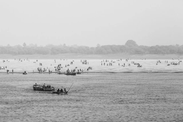 Barcos num rio e muitas pessoas na margem oposta, fotografados a preto e branco na Índia.