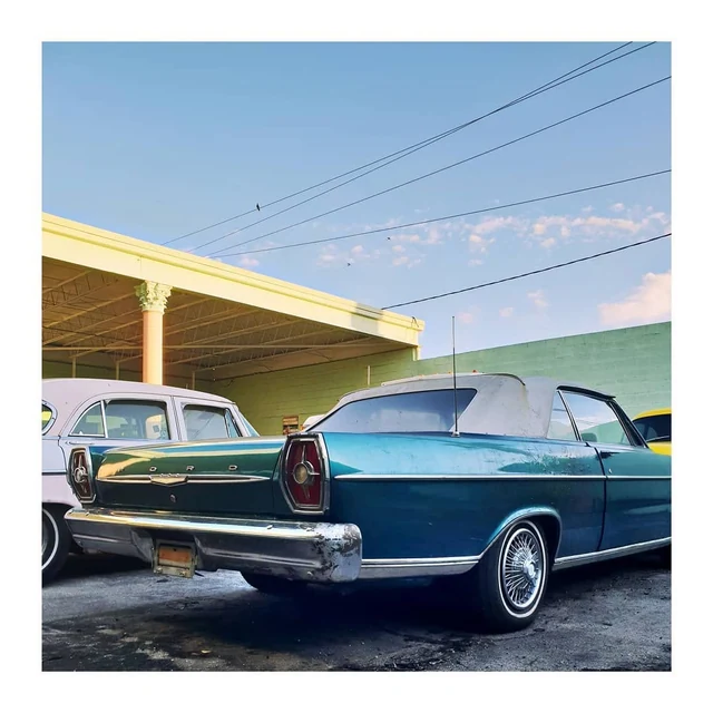 Um automóvel antigo azul-turquesa e branco diante de uma oficina com paredes em tons pastel, sob um céu atravessado por cabos.
